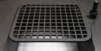 A square black cast iron grate cover with a grid pattern, shown installed on a metal surface with a small black knob visible on the right side.