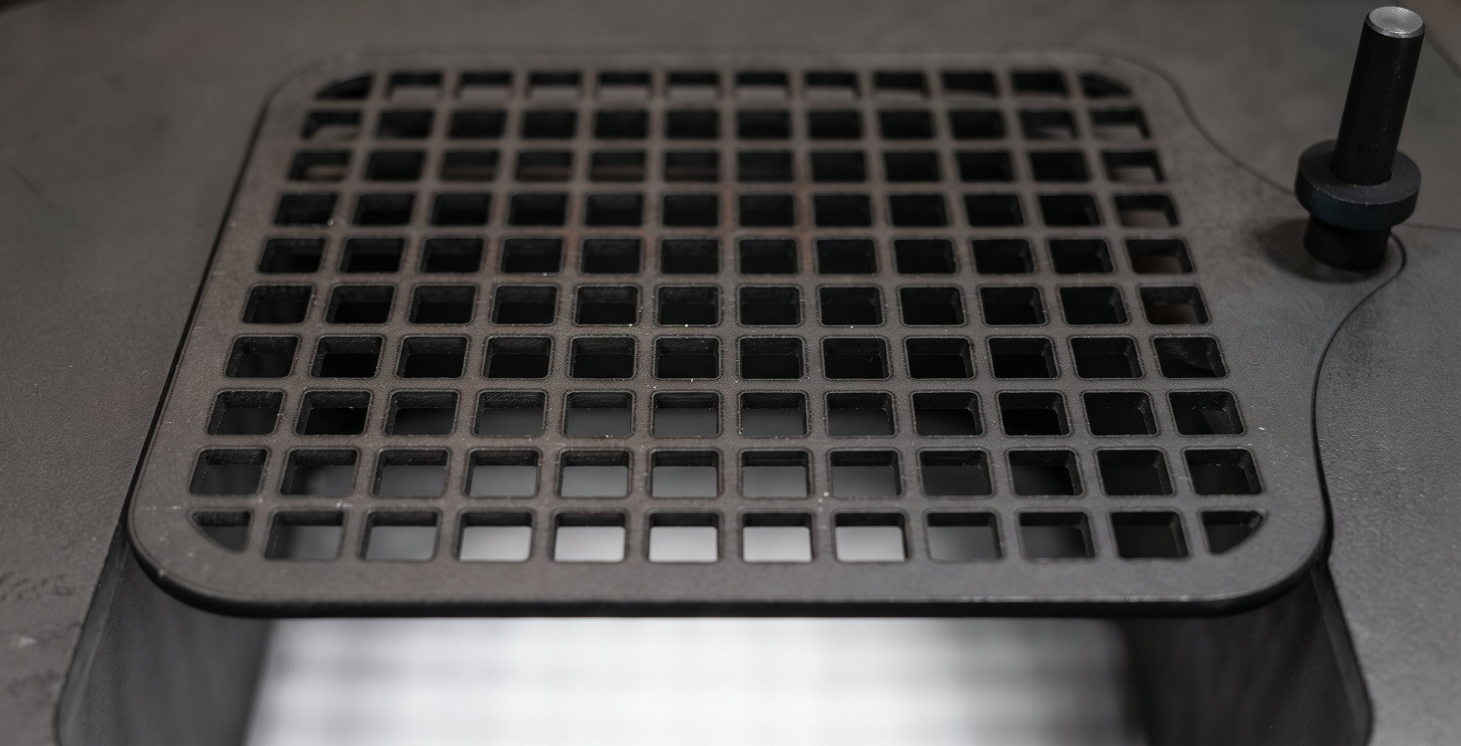 A square black cast iron grate cover with a grid pattern, shown installed on a metal surface with a small black knob visible on the right side.