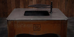 A Black Earth outdoor fire pit table with a rusted brown base, a flat metal top, a black firebox opening, and a hanging black grate, shown against a dark wood plank background.