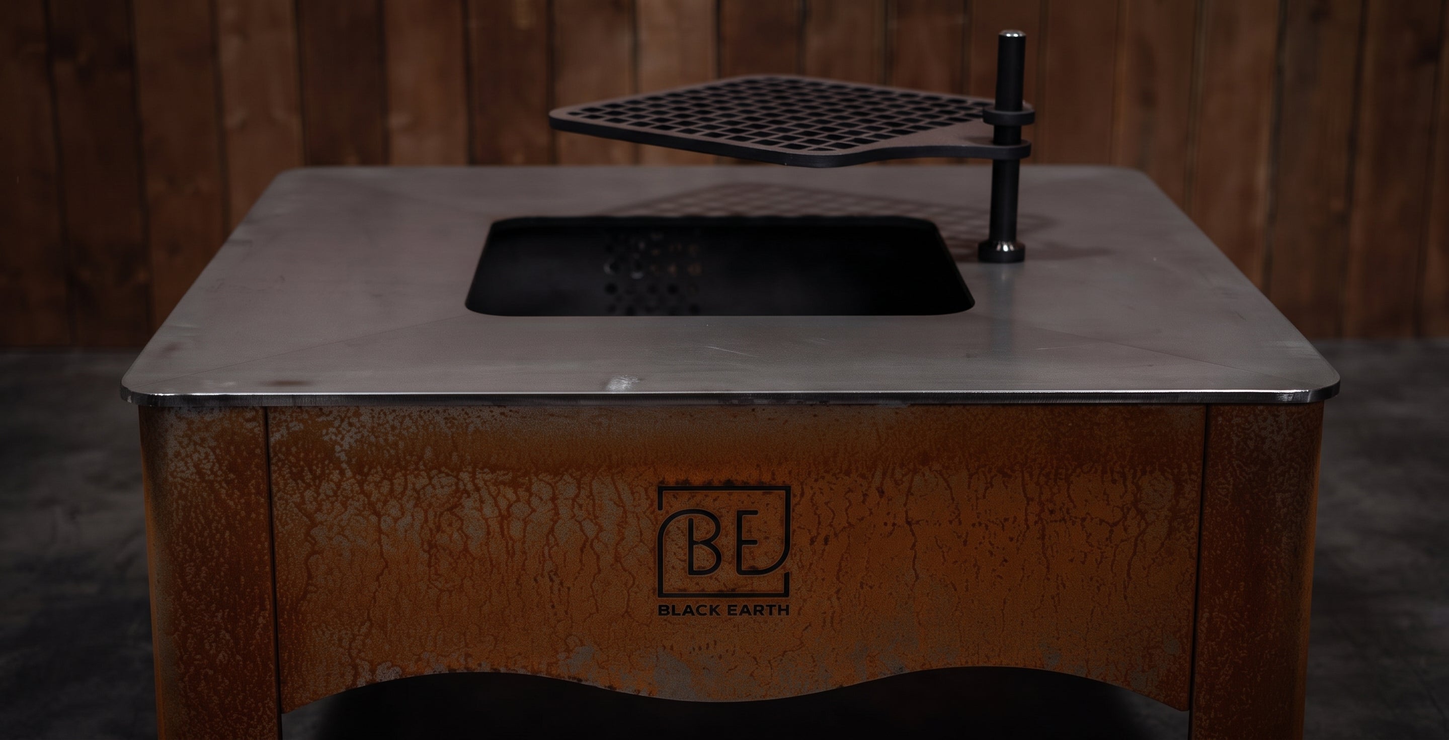 A Black Earth outdoor fire pit table with a rusted brown base, a flat metal top, a black firebox opening, and a hanging black grate, shown against a dark wood plank background.