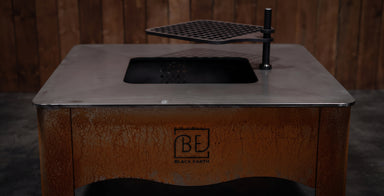 A Black Earth outdoor fire pit table with a rusted brown base, a flat metal top, a black firebox opening, and a hanging black grate, shown against a dark wood plank background.